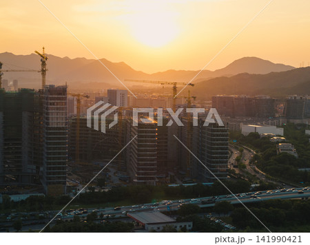 Tall residential buildings under construction with multiple cranes rising above the city skyline during a golden sunset near mountains in China Tall residential buildings under construction with multiple cranes rising above the city skyline during a golden sunset near mountains in China 141990421