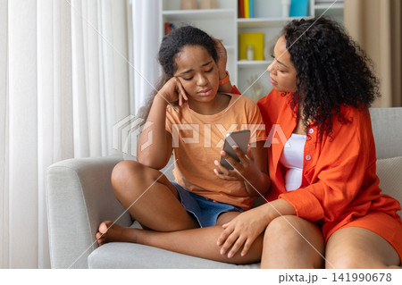 Concerned Black mother comforting her upset daughter while she looking at smartphone Concerned Black mother comforting her upset daughter while she looking at smartphone 141990678