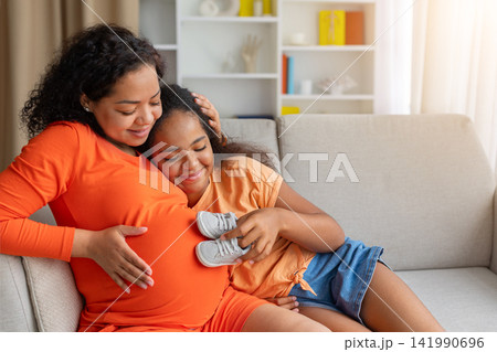 Black mother and daughter cuddling on sofa while holding tiny baby shoes Black mother and daughter cuddling on sofa while holding tiny baby shoes 141990696