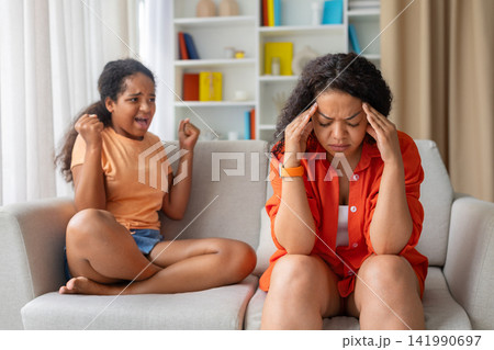 Stressed black mother holding head in pain while daughter shouts angrily behind 141990697