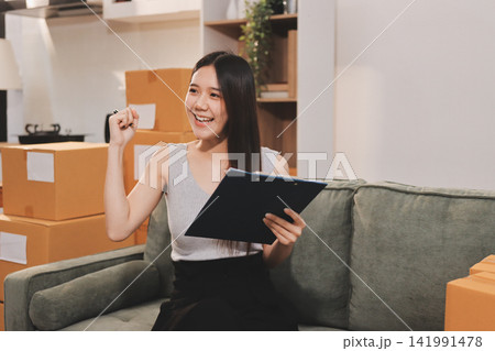 Startup SME small business entrepreneur of freelance Asian woman wearing apron using laptop and box to receive and review orders online to prepare to pack sell to customers, online sme business ideas. Startup SME small business entrepreneur of freelance Asian woman wearing apron using laptop and box to receive and review orders online to prepare to pack sell to customers, online sme business ideas. 141991478