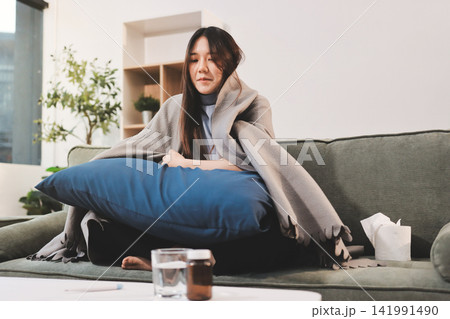 sick asian young woman sneezing into tissue paper while covered with a blanket and sitting on sofa in the living room 141991490