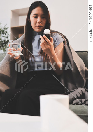 sick asian young woman sneezing into tissue paper while covered with a blanket and sitting on sofa in the living room 141991495