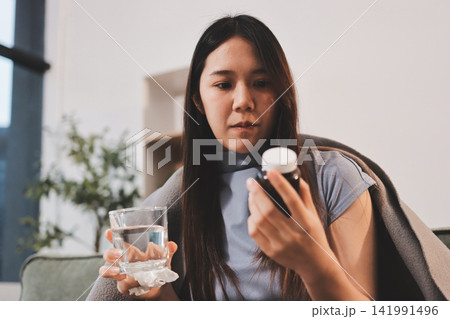 sick asian young woman sneezing into tissue paper while covered with a blanket and sitting on sofa in the living room sick asian young woman sneezing into tissue paper while covered with a blanket and sitting on sofa in the living room 141991496