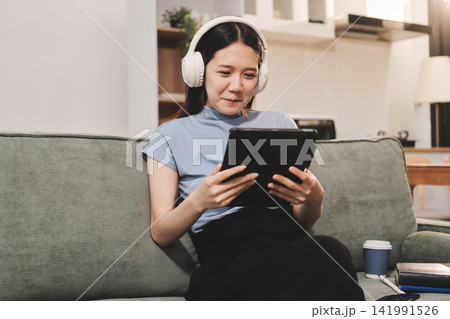 Freelance Asian woman holding mobile phone typing on laptop and working from home. Happy young girl sitting on sofa in living room. Freelance Asian woman holding mobile phone typing on laptop and working from home. Happy young girl sitting on sofa in living room. 141991526