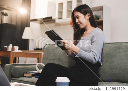 Freelance Asian woman holding mobile phone typing on laptop and working from home. Happy young girl sitting on sofa in living room. Freelance Asian woman holding mobile phone typing on laptop and working from home. Happy young girl sitting on sofa in living room. 141991529