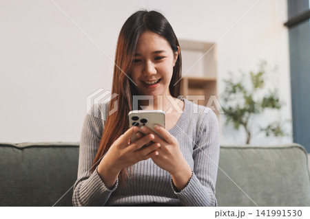 Happy relaxed young woman sitting on couch using cell phone, smiling lady laughing holding smartphone, looking at cellphone enjoying doing online ecommerce shopping in mobile apps or watching videos. 141991530