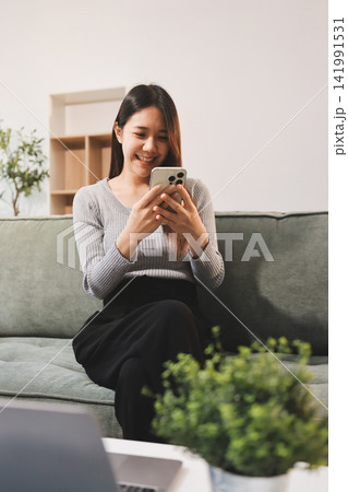 Happy relaxed young woman sitting on couch using cell phone, smiling lady laughing holding smartphone, looking at cellphone enjoying doing online ecommerce shopping in mobile apps or watching videos. Happy relaxed young woman sitting on couch using cell phone, smiling lady laughing holding smartphone, looking at cellphone enjoying doing online ecommerce shopping in mobile apps or watching videos. 141991531