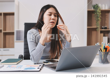 Young Asian woman sits at office desk rubbings her eyes and temples feeling pain. Stress. And exhaustion from long hours working on the computer 141991545