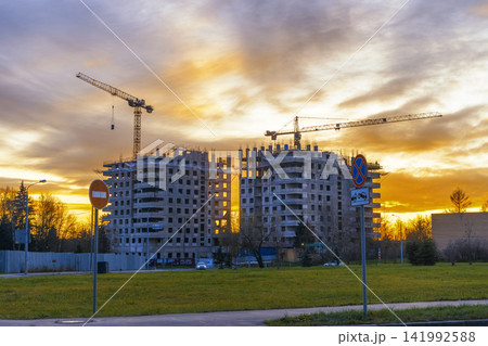 Unfinished concrete apartment blocks with tower cranes against dramatic golden sunset sky Unfinished concrete apartment blocks with tower cranes against dramatic golden sunset sky 141992588