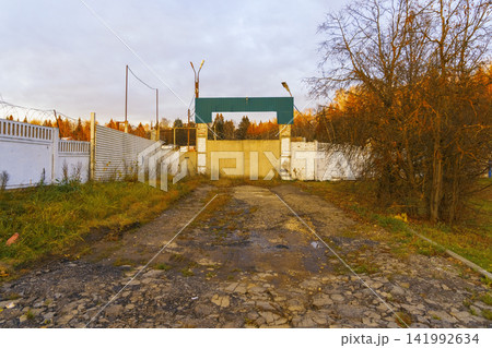 Rundown gated entrance with cracked driveway, tall fence panels, and overgrown verge at dusk Rundown gated entrance with cracked driveway, tall fence panels, and overgrown verge at dusk 141992634