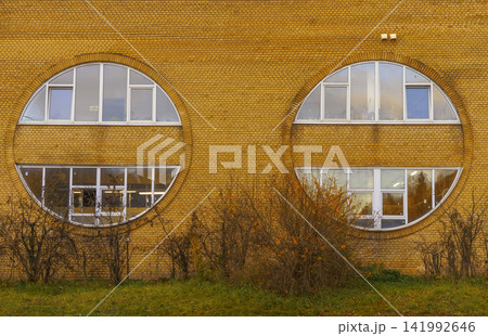Yellow brick building facade with two large round windows, white frames, and neatly trimmed shrubs Yellow brick building facade with two large round windows, white frames, and neatly trimmed shrubs 141992646