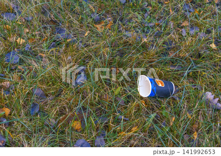 Discarded paper cup lying in grassy field with autumn leaves, showing outdoor litter and seasonal neglect Discarded paper cup lying in grassy field with autumn leaves, showing outdoor litter and seasonal neglect 141992653