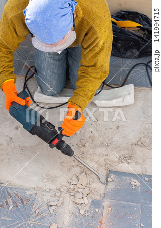 Person in orange gloves and work clothes uses a hammer drill to chip old grey tiles from an open apartment balcony floor. The tool breaks the surface, creating dust and tile debris Person in orange gloves and work clothes uses a hammer drill to chip old grey tiles from an open apartment balcony floor. The tool breaks the surface, creating dust and tile debris 141994715