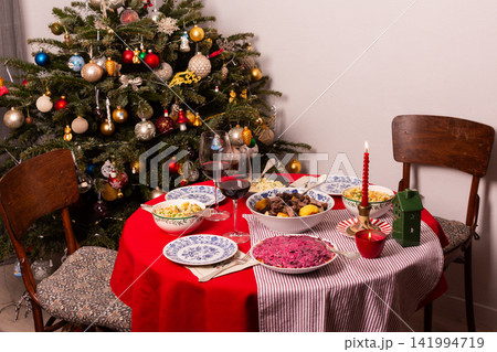 A table set for a Christmas and New Year celebration displays various dishes like salad, meat, and grains. A red candle, wine glass, and colorful decorations add to the festive atmosphere 141994719