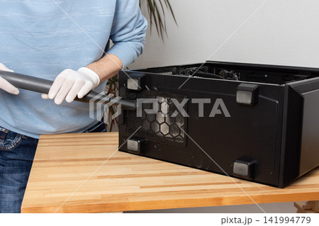 A 53-year-old man uses a vacuum to clean dust from a computer system unit on a wooden table in a home office. The setting is tidy and organized 141994779