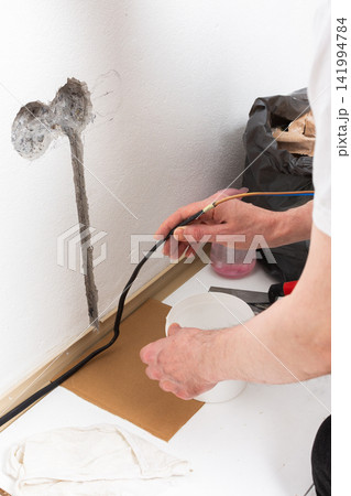 Worker runs new electrical wires into a cutout in a white apartment wall. This task prepares for the installation of an electrical outlet near the baseboard 141994784