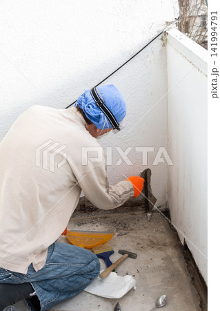 Person wearing blue head covering, safety goggles, and orange gloves applies cement to an apartment balcony wall. Renovation work improves the open outdoor space, focusing on corner repairs 141994791