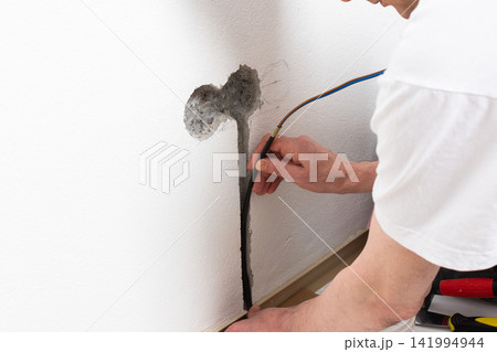 Worker runs new electrical wires into a cutout in a white apartment wall. This task prepares for the installation of an electrical outlet near the baseboard Worker runs new electrical wires into a cutout in a white apartment wall. This task prepares for the installation of an electrical outlet near the baseboard 141994944