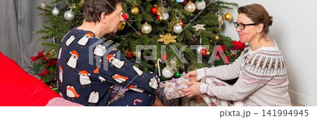 A couple sits near a Christmas tree on Christmas Day. They are exchanging gifts and enjoying the holiday spirit. The room has decorations and a festive atmosphere A couple sits near a Christmas tree on Christmas Day. They are exchanging gifts and enjoying the holiday spirit. The room has decorations and a festive atmosphere 141994945