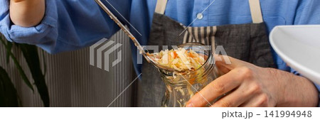 A 53-year-old man works in his kitchen fermenting cabbage and carrots. He carefully adds the mixture from a large bowl into a jar, showing concentration, banner 141994948