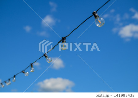Clear glass bulbs hang from a wire against a bright blue sky. White clouds float gently in the background. Each bulb is simple, unlit, and slightly frosted 141995438