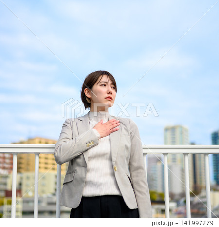 屋上で遠くを見つめながら胸に手を当てる20代の日本人女性 屋上で遠くを見つめながら胸に手を当てる20代の日本人女性 141997279