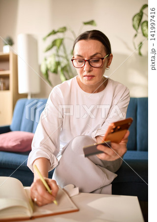 Mature woman confirming online transfer while taking notes at home Mature woman confirming online transfer while taking notes at home 141997416