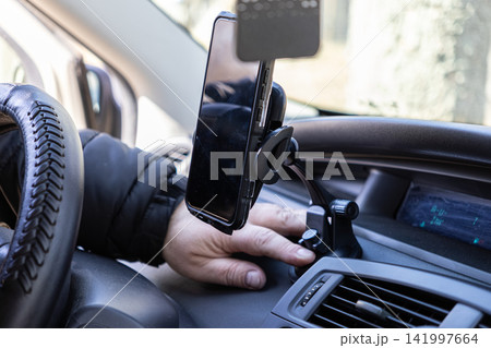 A person adjusts a smartphone mounted on a car dashboard holder for safe navigation during travel, illustrating modern connectivity and hands-free driving assistance in a vehicle A person adjusts a smartphone mounted on a car dashboard holder for safe navigation during travel, illustrating modern connectivity and hands-free driving assistance in a vehicle 141997664
