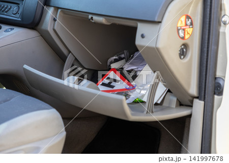 Open glove compartment of a vehicle containing various scattered items and a student driver H sign, capturing an everyday automotive storage mess in a practical travel setup 141997678