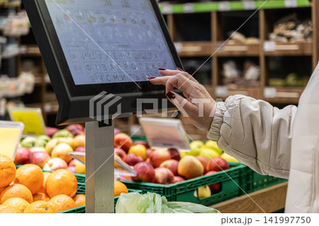 A customer uses a modern touchscreen scale to weigh fresh fruit in a busy grocery supermarket, highlighting self-service retail efficiency and a seamless daily shopping experience 141997750