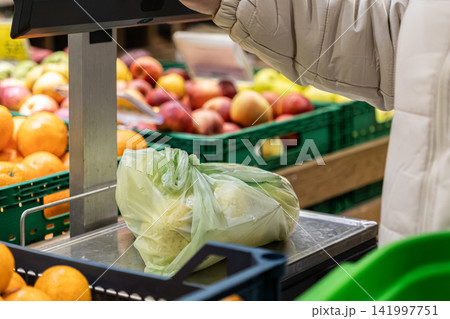 A person carefully weighs a plastic bag of fresh produce on a digital supermarket scale, highlighting healthy shopping habits and daily food consumption in a vibrant grocery store 141997751