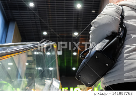 Person in a white jacket carrying a black leather bag stands near a shopping mall escalator, depicting daily urban life, consumer travel, and modern transit in a commercial setting 141997768