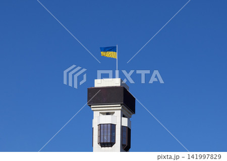 The Ukrainian flag flutters proudly atop a white architectural tower against a vibrant clear blue sky, symbolizing national independence and resilience in a bright urban landscape The Ukrainian flag flutters proudly atop a white architectural tower against a vibrant clear blue sky, symbolizing national independence and resilience in a bright urban landscape 141997829
