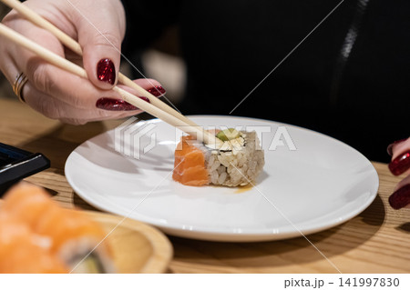 Person using wooden chopsticks to pick up a single piece of fresh salmon sushi roll from a white plate in a restaurant setting, highlighting culinary enjoyment and Japanese cuisine Person using wooden chopsticks to pick up a single piece of fresh salmon sushi roll from a white plate in a restaurant setting, highlighting culinary enjoyment and Japanese cuisine 141997830