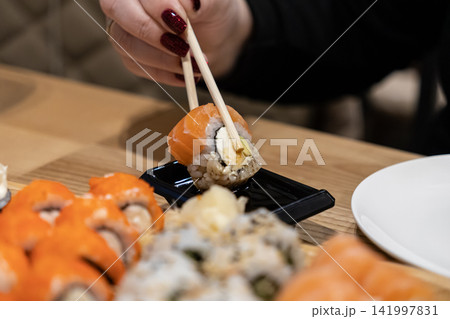 Woman picking up a single fresh salmon sushi roll with wooden chopsticks to dip into a black soy sauce container, representing authentic Japanese dining and healthy gourmet cuisine Woman picking up a single fresh salmon sushi roll with wooden chopsticks to dip into a black soy sauce container, representing authentic Japanese dining and healthy gourmet cuisine 141997831