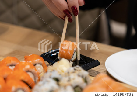 Person with red manicured nails holding a salmon sushi roll with chopsticks, dipping it into soy sauce at a restaurant table to highlight a Japanese fine dining culinary experience Person with red manicured nails holding a salmon sushi roll with chopsticks, dipping it into soy sauce at a restaurant table to highlight a Japanese fine dining culinary experience 141997832