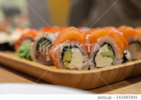 Fresh salmon sushi rolls with cream cheese and avocado arranged on a wooden serving tray, evoking a premium dining experience at a restaurant or a relaxed social gathering meal Fresh salmon sushi rolls with cream cheese and avocado arranged on a wooden serving tray, evoking a premium dining experience at a restaurant or a relaxed social gathering meal 141997845