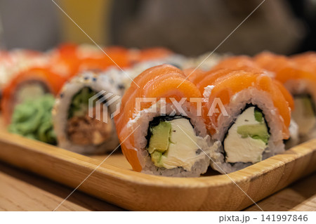 Fresh salmon sushi rolls filled with avocado and cream cheese on a bamboo serving tray, creating an appealing presentation for Japanese cuisine dining in a modern restaurant setup Fresh salmon sushi rolls filled with avocado and cream cheese on a bamboo serving tray, creating an appealing presentation for Japanese cuisine dining in a modern restaurant setup 141997846