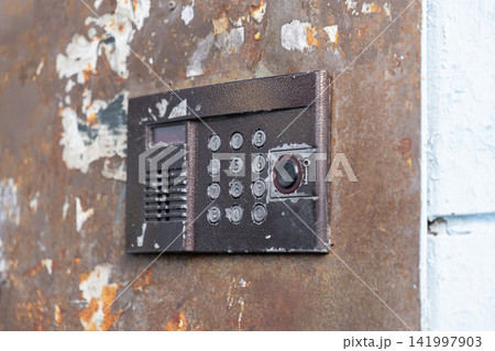 A worn intercom keypad is mounted on a decaying rusty metal door, representing urban decay and architectural neglect while highlighting security hardware in a gritty city setting A worn intercom keypad is mounted on a decaying rusty metal door, representing urban decay and architectural neglect while highlighting security hardware in a gritty city setting 141997903