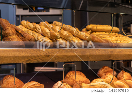Fresh baguettes and artisan bread loaves displayed on a rustic wooden bakery shelf, conveying a warm traditional atmosphere of a neighborhood boulangerie offering handmade goods 141997910