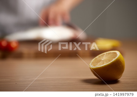 Lemon half in focus on a kitchen table, person preparing fresh fish and vegetables in blurred background, cooking seafood 141997979