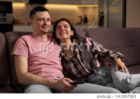 Happy couple enjoying a cozy evening controlling the tv with a remote, sharing leisure time on the sofa 141997981