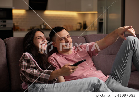 Happy couple relaxing on a sofa, watching tv and laughing together. woman holding remote control, pointing at screen 141997983