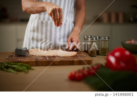 Chef's hands adding coarse salt to raw chicken fillets on a wooden cutting board, preparing a meal in the kitchen Chef's hands adding coarse salt to raw chicken fillets on a wooden cutting board, preparing a meal in the kitchen 141998001