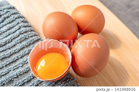 Brown chicken eggs and cracked egg with fresh yolk on wooden cutting board beside gray kitchen towel close up food photo Brown chicken eggs and cracked egg with fresh yolk on wooden cutting board beside gray kitchen towel close up food photo 141998016
