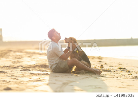 Man sitting on sandy beach with dog during sunset, engaging in playful interaction, ocean waves and distant pier visible in the background. Airedale Terrier Man sitting on sandy beach with dog during sunset, engaging in playful interaction, ocean waves and distant pier visible in the background. Airedale Terrier 141998038