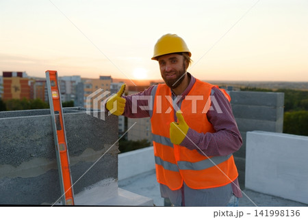 Construction worker wearing hardhat and safety vest leaning on concrete block at sunset 141998136