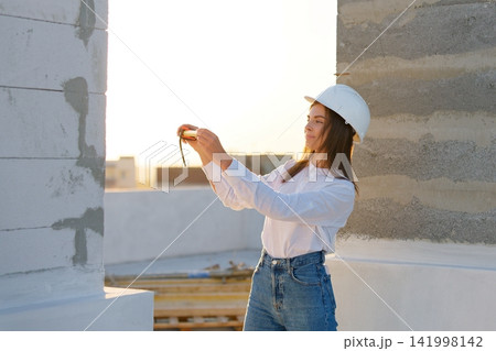 Female engineer measuring wall with tape measure at construction site during sunset 141998142