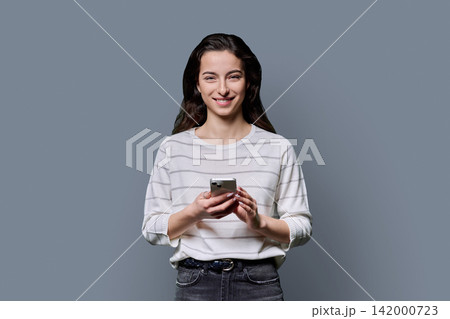 Smiling young woman holding smartphone and looking at camera over grey background Smiling young woman holding smartphone and looking at camera over grey background 142000723